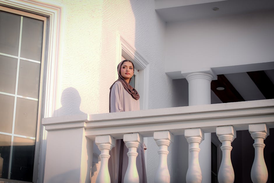 Woman in modest fashion standing on a sunlit balcony, exuding elegance and grace.